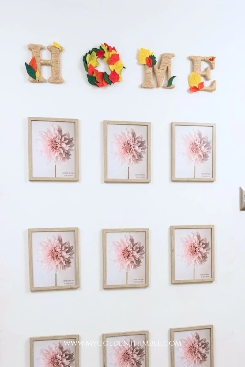 A vertical indoor wall shot illuminated with bright, clear morning natural light. The top wall decor spells "HOME" using twine-wrapped letters decorated with colorful red, yellow, and green felt leaves, including an autumn leaf wreath for the "O". Beneath the word, nine matching wood-framed pink dahlia botanical prints are displayed in a clean 3x3 grid above the staircase trim.