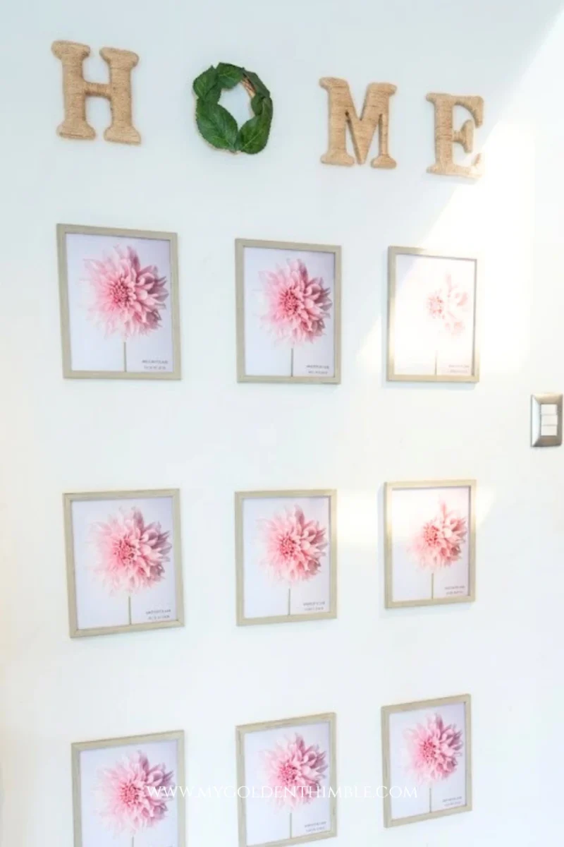 A full vertical shot of an indoor gallery wall lit by bright, clean morning sunlight coming from the side. At the top, twine-wrapped letters "H", "M", and "E" flank a small green leaf wreath for the "O". Below the spelling, nine matching wooden picture frames featuring pink dahlia floral prints are arranged in a neat 3x3 grid.