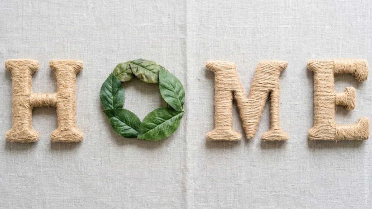 twine-wrapped letters spelling "HOME" arranged on a light-colored linen textured background. The letter "O" is replaced by a small, round green leaf wreath. Bright, soft morning sunlight casts gentle highlights across the letters.