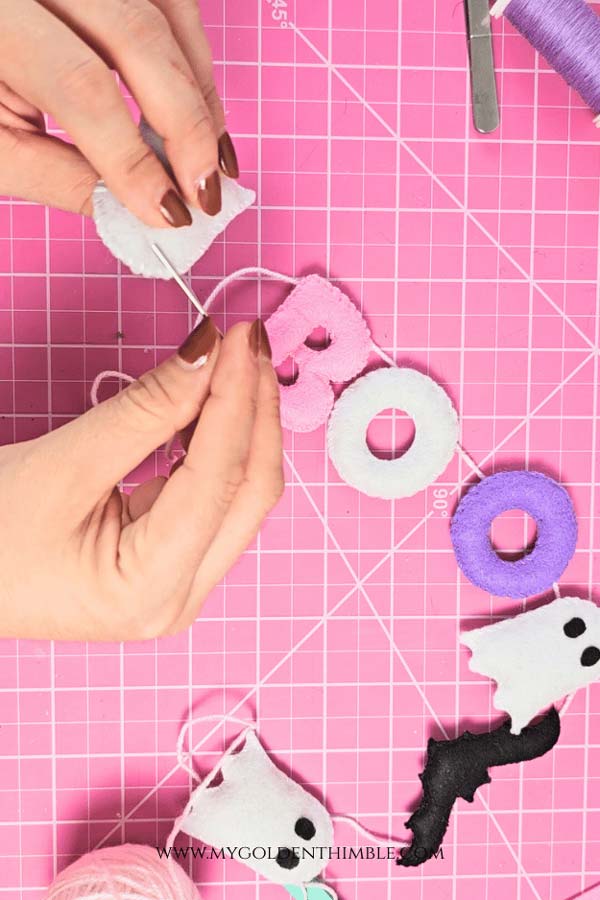 Assembling DIY Halloween garland, threading pink and purple felt letters, ghosts, and bats onto cord for hanging.
