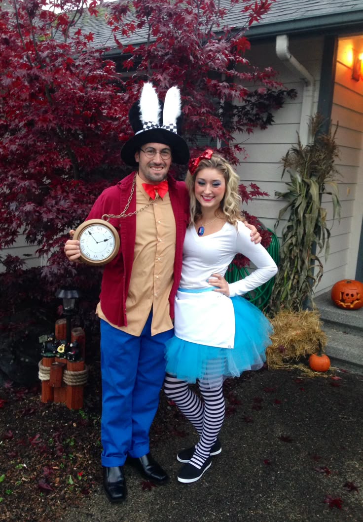 Two people dressed in costumes. The person on the left is wearing a red coat, blue pants, and a top hat with rabbit ears, holding a large pocket watch. Costume ideas for adults