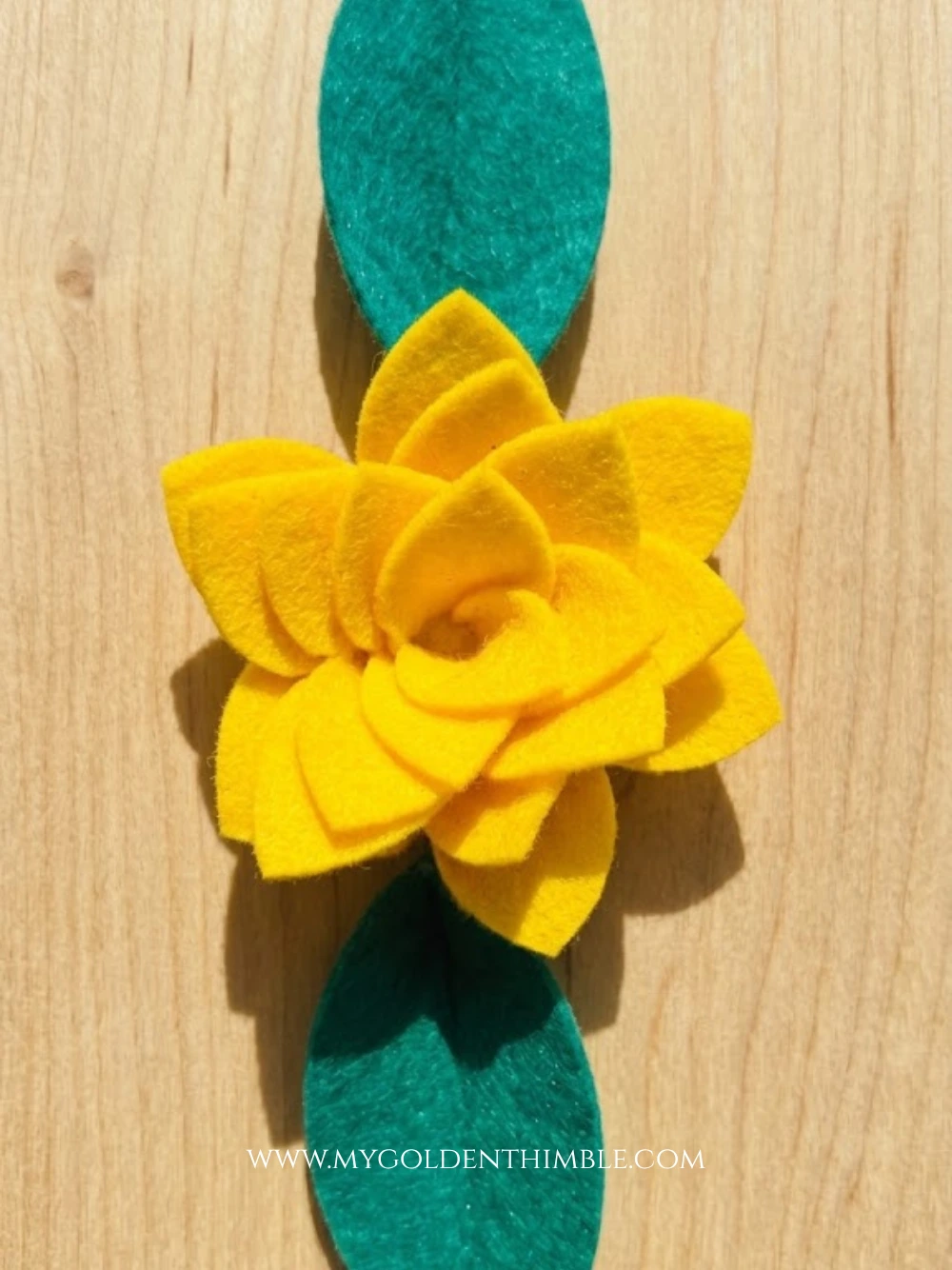 Close-up flat lay of a handmade bright yellow felt layered flower with two teal-green felt leaves extended from top and bottom, set on a light wooden background.