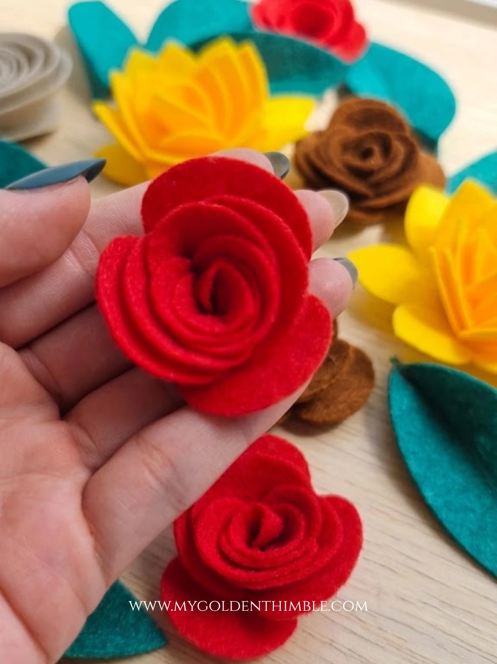 Hand holding a vibrant red rolled felt rose over a wooden workspace surrounded by additional yellow, brown, and red felt flowers and green leaves.