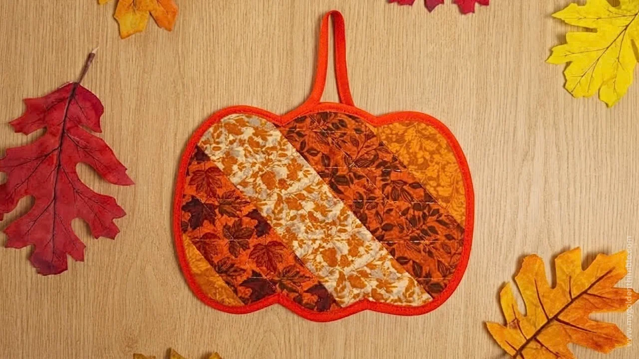 Finished pumpkin pot holder in warm burnt orange and fall-colored fabrics displayed on a wooden table with decorative leaves.