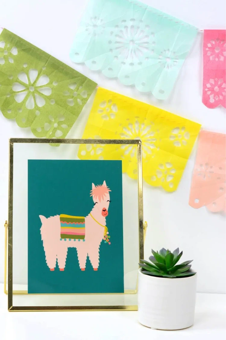 Framed llama wall art displayed on a desk next to a potted succulent with pastel papel picado banners.