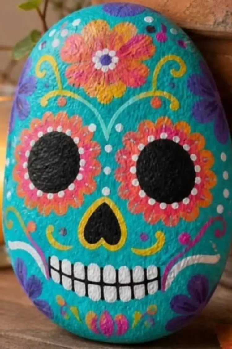 Hand-painted oval rock decorated as a turquoise sugar skull with colorful floral patterns.
