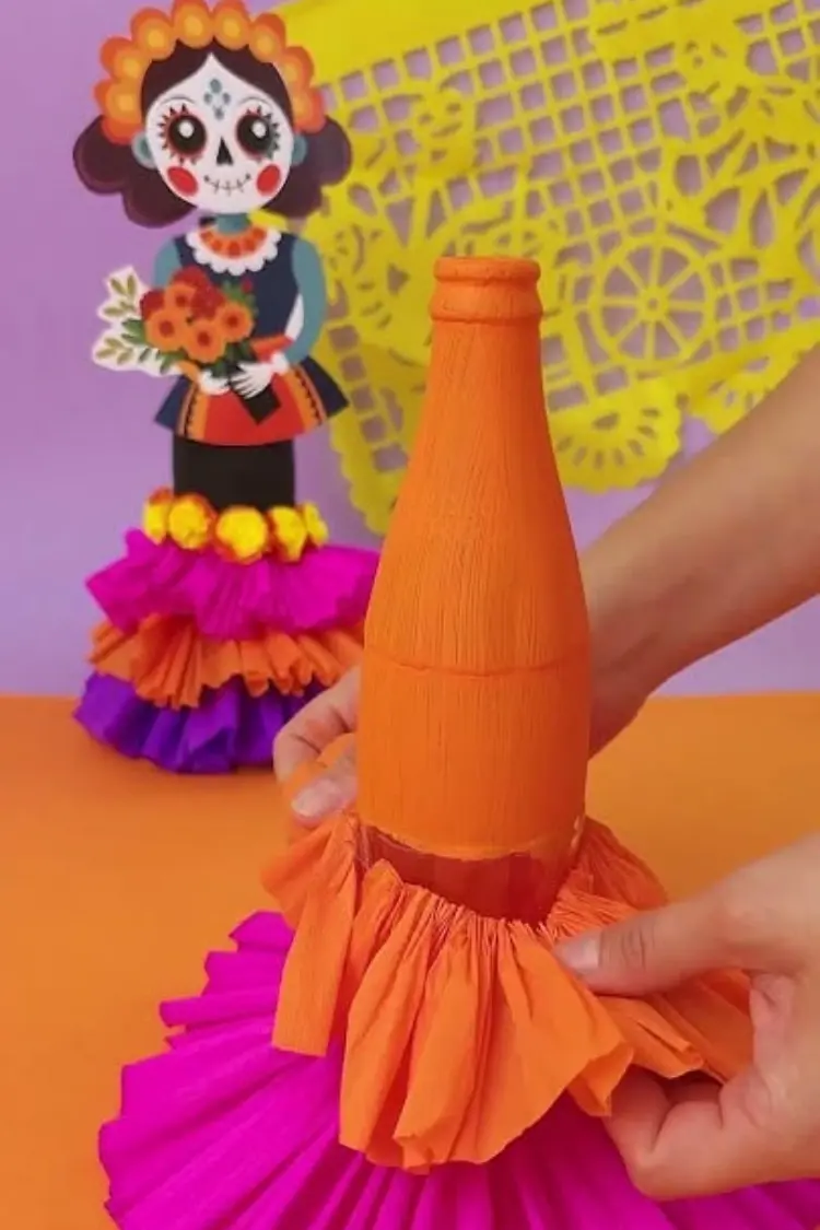 Hands wrapping bright orange tissue paper around a glass bottle to make a Catrina figure craft.