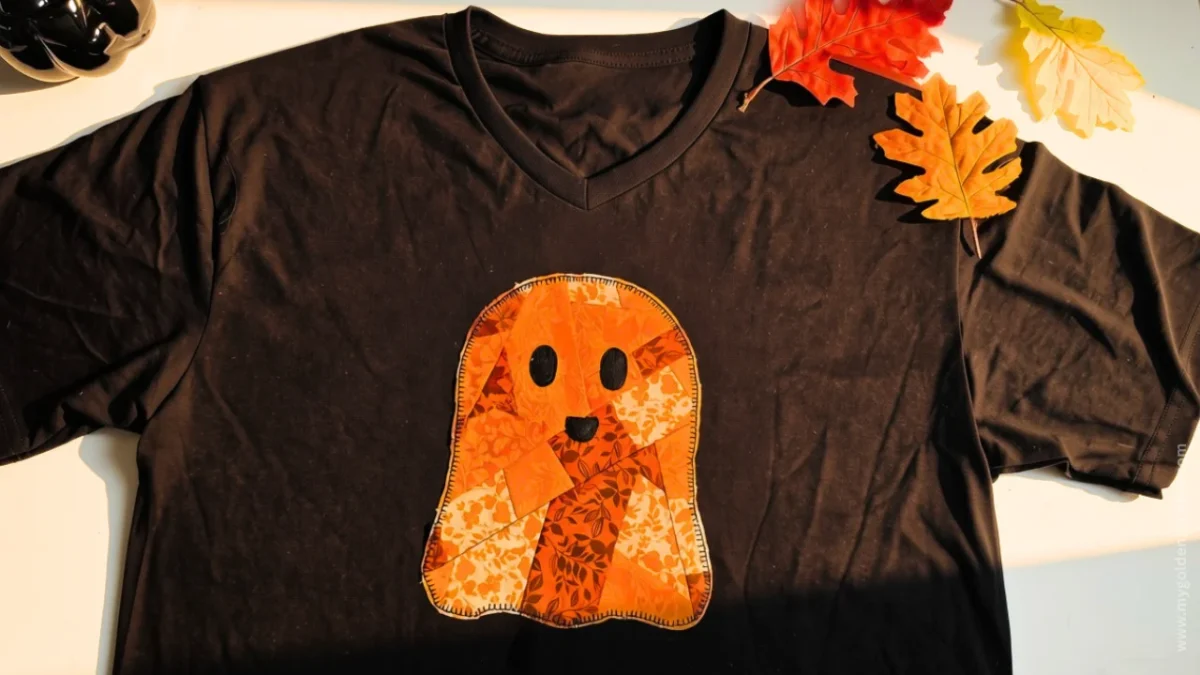 Horizontal flat lay of a black V-neck t-shirt with an orange patchwork ghost appliqué and fall leaves on a wooden surface.