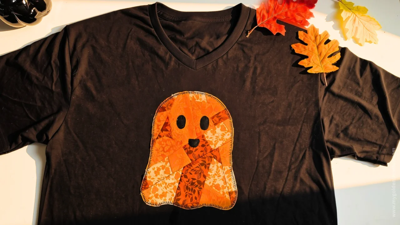 Horizontal flat lay of a black V-neck t-shirt with an orange patchwork ghost appliqué and fall leaves on a wooden surface.