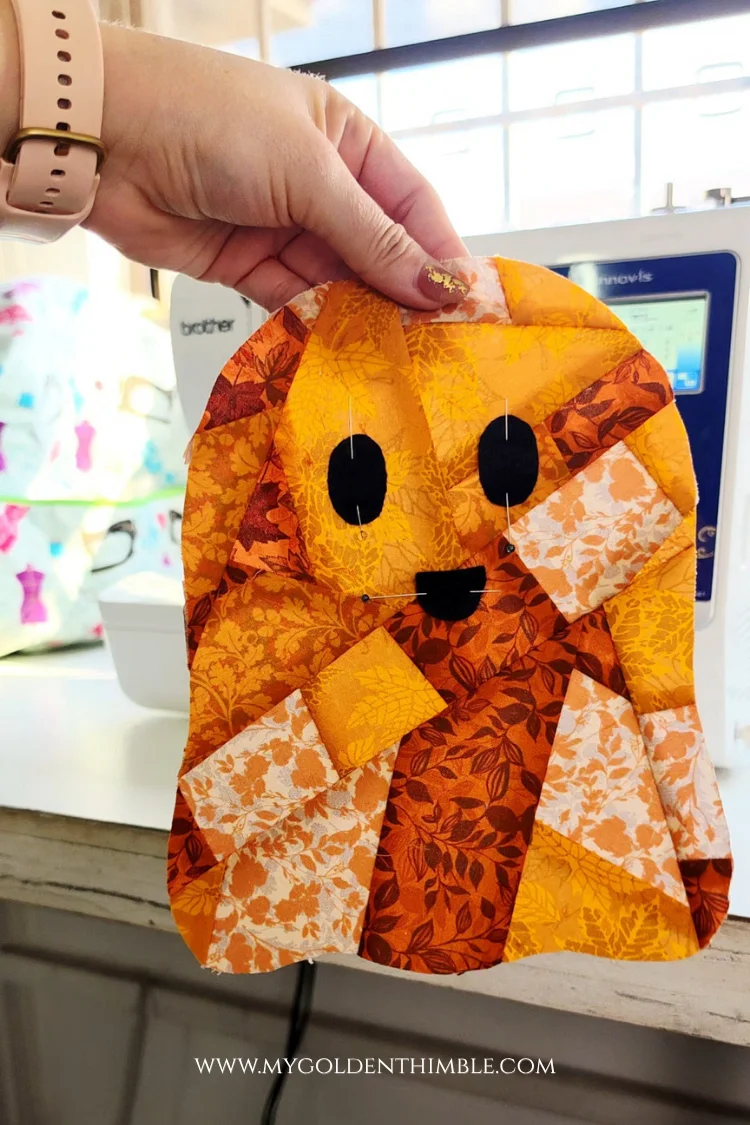 Hands holding up a finished orange patchwork ghost appliqué with embroidered eyes and mouth.