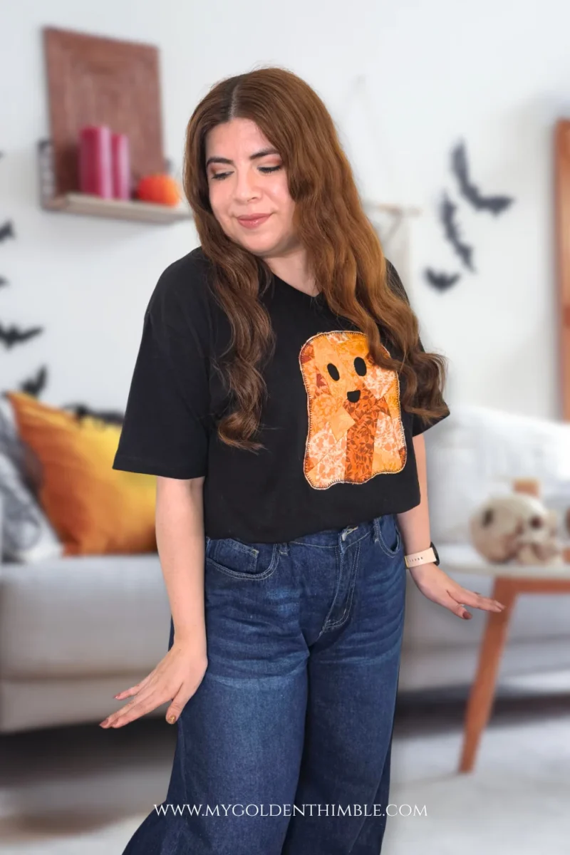 Woman wearing a black t-shirt featuring a DIY orange patchwork ghost appliqué in a Halloween-decorated living room.