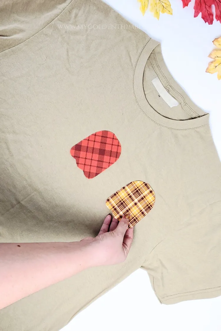 Hand placing plaid ghost fabric appliques onto the chest area of a beige t-shirt.
