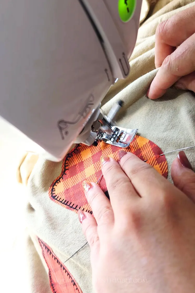 Close-up of hands guiding a beige shirt under a sewing machine presser foot to stitch down a plaid ghost applique.