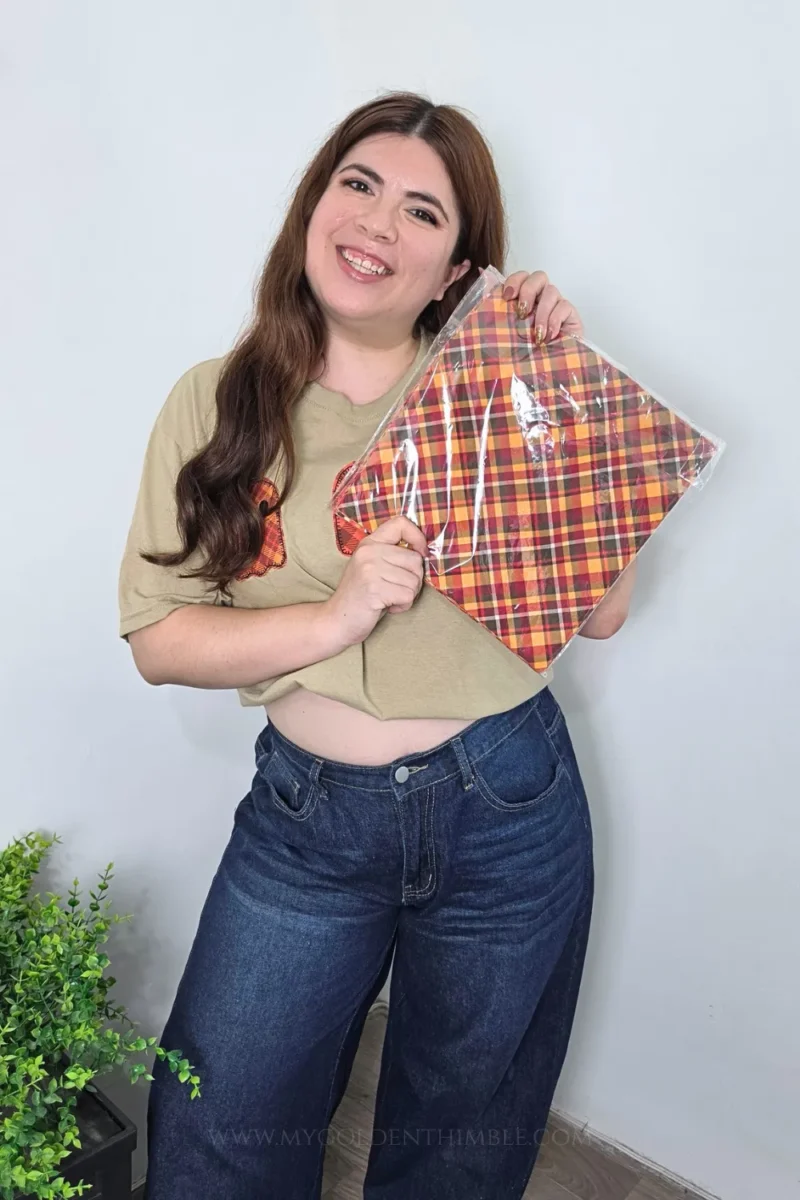 A woman smiling while holding plaid fabric sheets, wearing the completed beige t-shirt with three ghost appliques and dark blue jeans.
