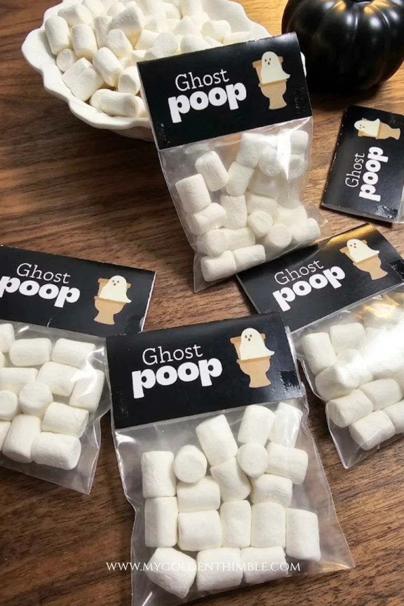 Flat lay of multiple clear treat bags filled with white mini marshmallows, topped with black folded tags reading 'Ghost Poop', displayed on a dark wood surface.