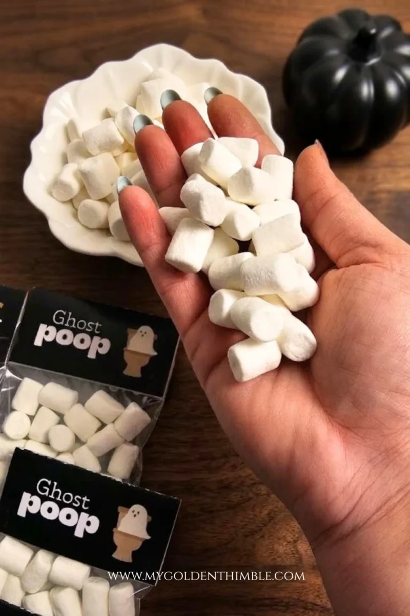 A hand holding a handful of loose white mini marshmallows over a wooden table styled with 'Ghost Poop' Halloween treat bags and a decorative black pumpkin